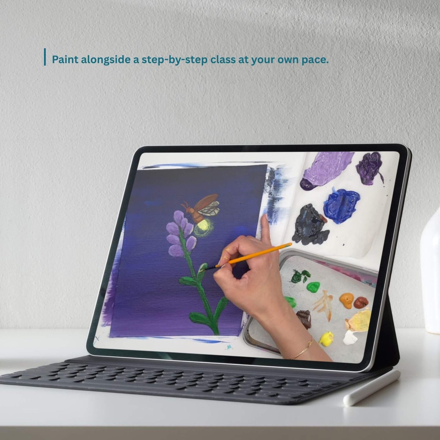 A tablet in a black protective case with a keyboard cover stands on a white surface against a light gray wall. The screen shows a hand holding a paintbrush and painting alongside a digital paint palette.