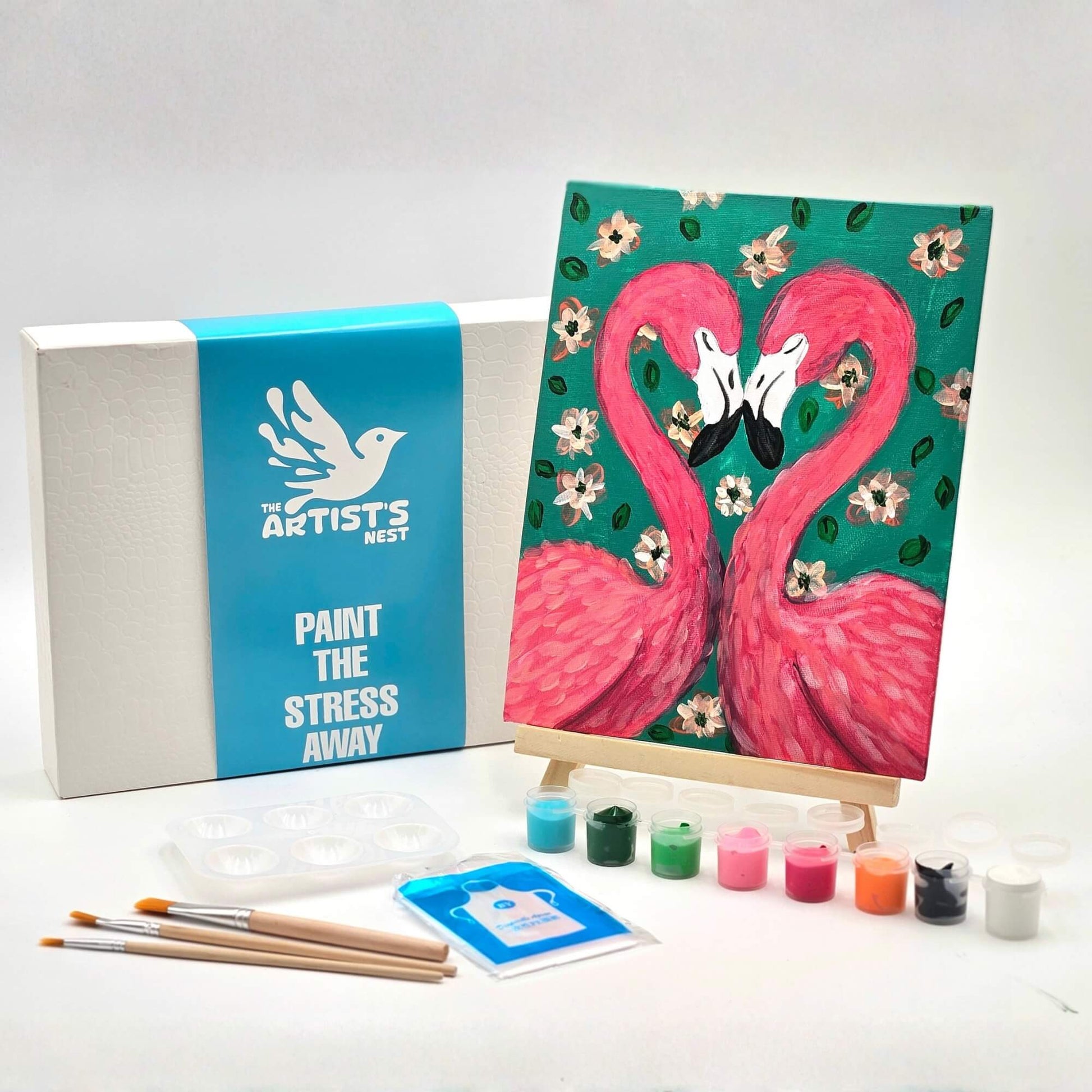 Art kit with a completed painting of two pink flamingos on a teal background, surrounded by paint bottles and brushes.