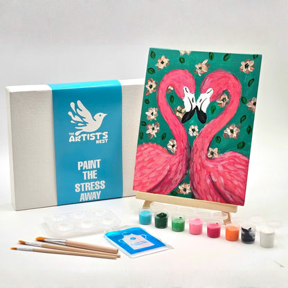 Art kit with a completed painting of two pink flamingos on a teal background, surrounded by paint bottles and brushes.