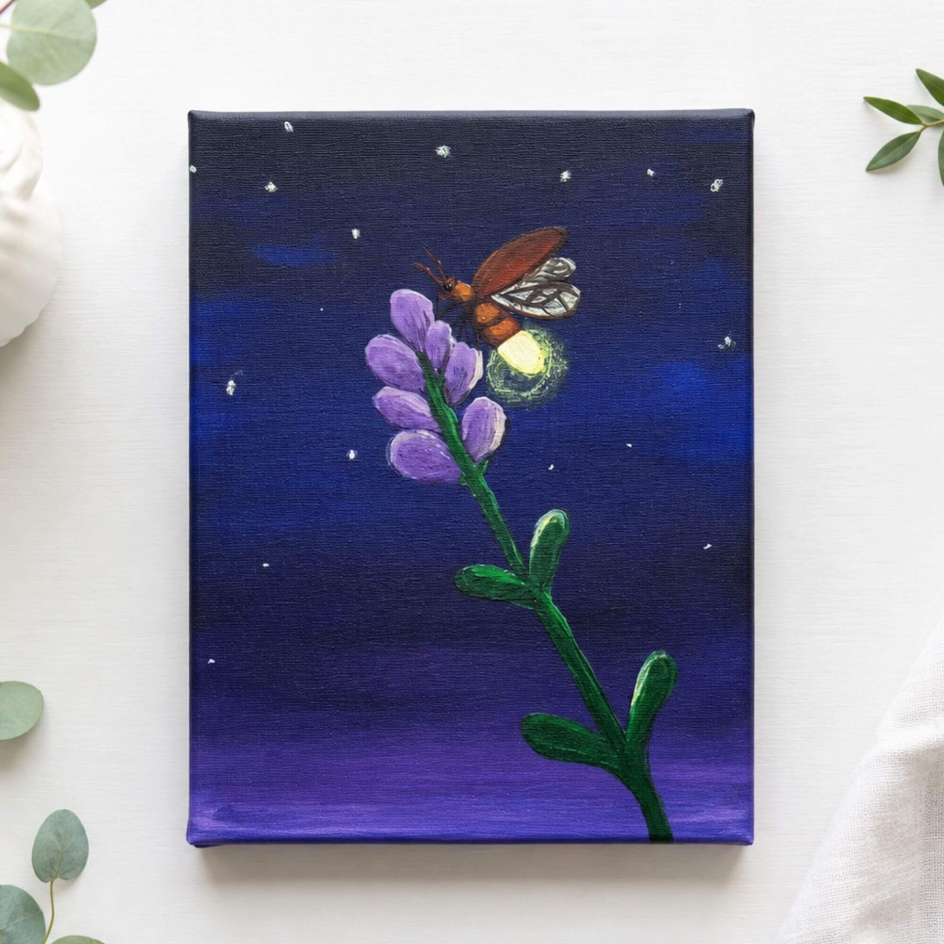 A square canvas painting featuring a glowing firefly on a purple flower stem against a deep blue and purple night sky, styled on a white surface with green leaves and a white vase in the background.