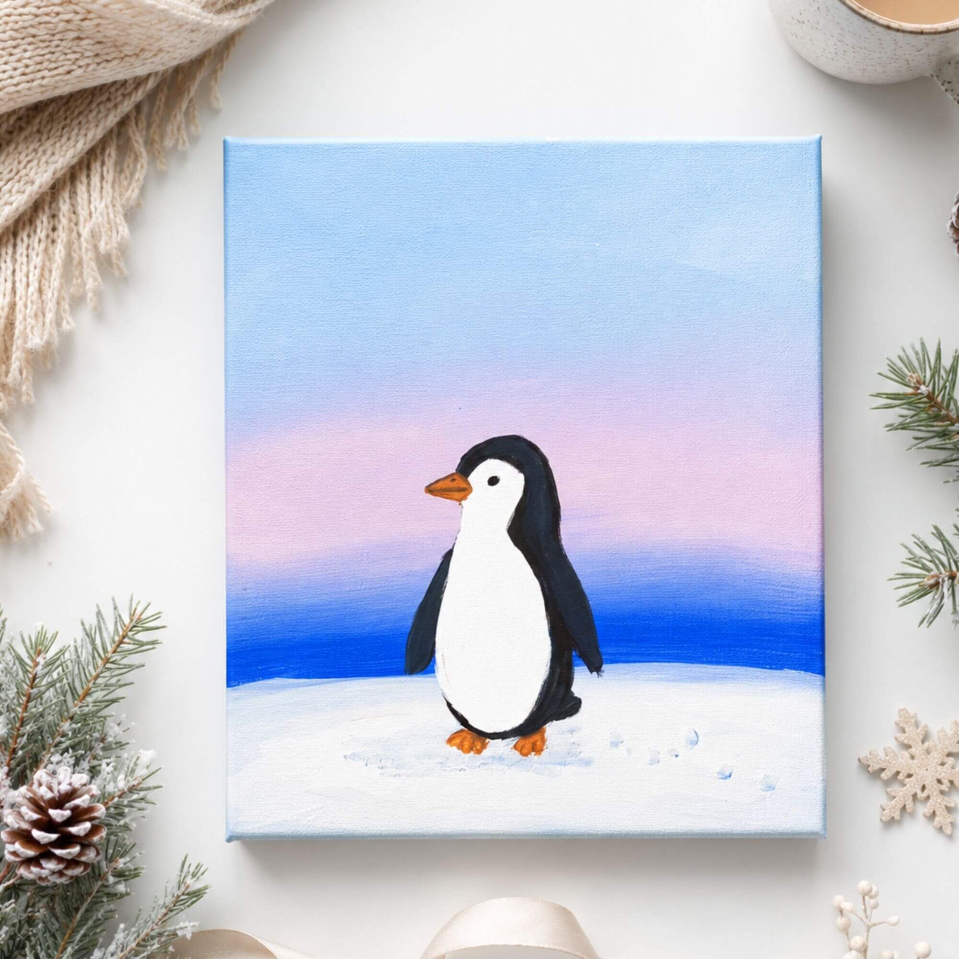A square canvas painting featuring a cute penguin standing on snowy ground under a pastel sky, styled on a white surface with holiday greenery, a scarf, and a mug around it.