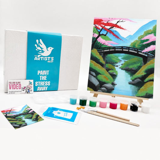 Paint kit by The Artists Nest, featuring a canvas print of a scenic landscape with a bridge and river, a white box with a blue band, paint tubes, brushes, and a small instruction card on a white background.