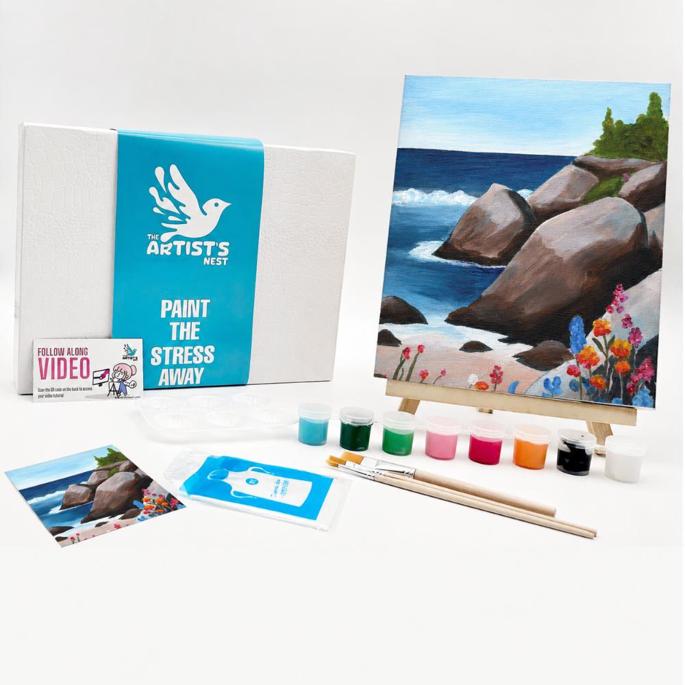 Painting kit set displayed on a white background, including a canvas box with a blue band reading 'The Artists Nest', a painted coastal scene on an easel, small paint pots, brushes, a printed photo, and a small card.