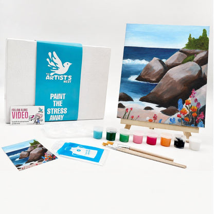Painting kit set displayed on a white background, including a canvas box with a blue band reading 'The Artists Nest', a painted coastal scene on an easel, small paint pots, brushes, a printed photo, and a small card.