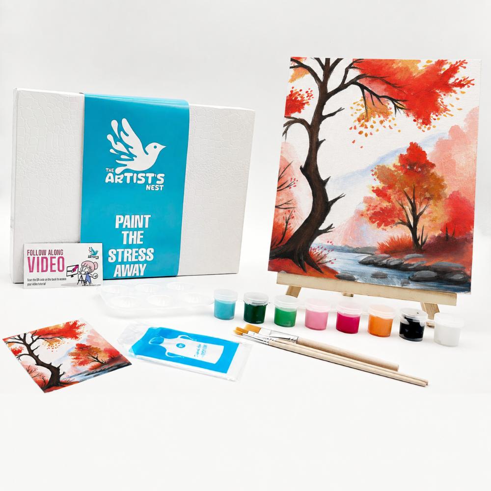 Painting kit set displayed on a white background, including a canvas box with a blue band, a painted autumn tree scene on a small easel, paint bottles, brushes, a small printed card, and a follow along video box.