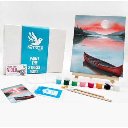 Painting kit set displayed on a white background, including a canvas painting of a red canoe on a lake at sunset, a white box with a blue band reading 'The Artists Nest', a small instruction card, paintbrushes, multiple paint pots, and a printed example painting.