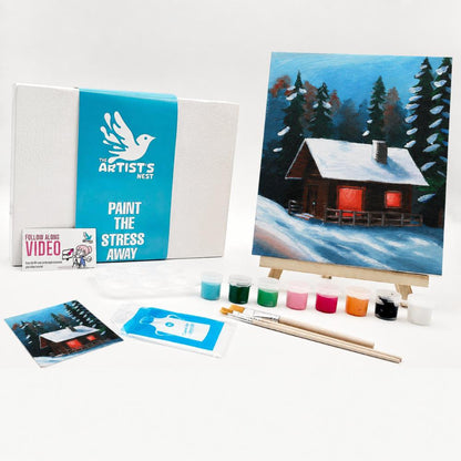 Paint kit by The Artists Nest, featuring a canvas print of a snowy cabin, a white and blue box, paint tubes, brushes, and instruction cards on a white background.